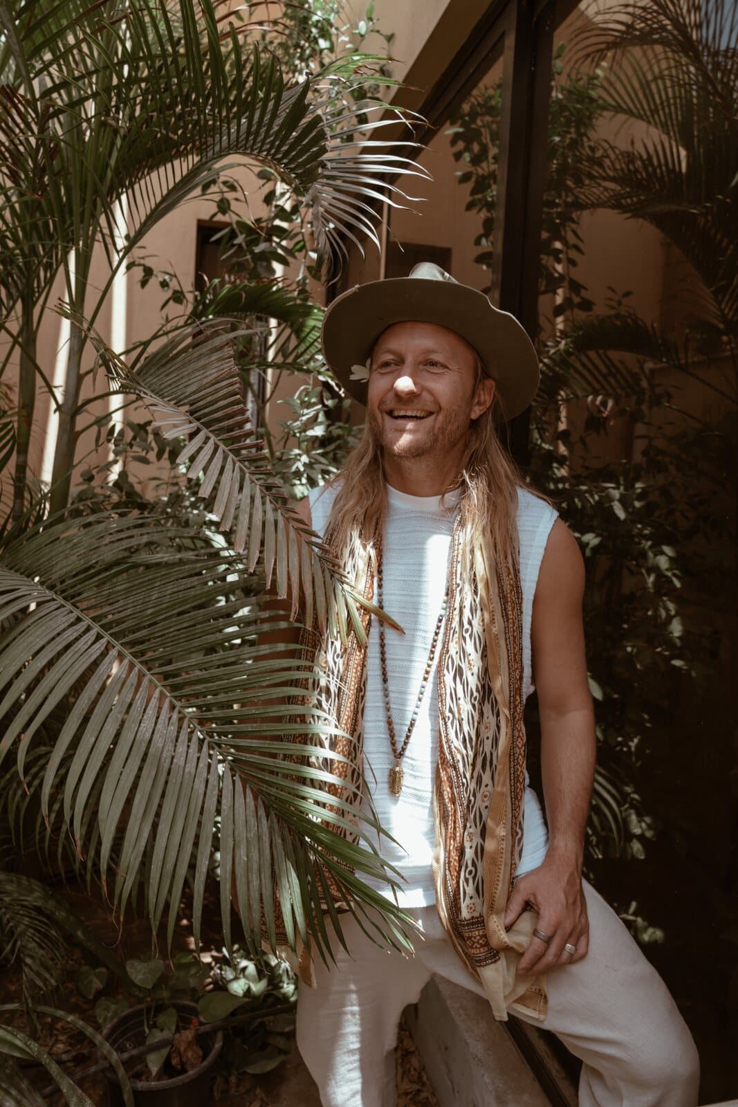 Victor Mathys laughing among palm fronds in a wide-brim hat, one hand raised